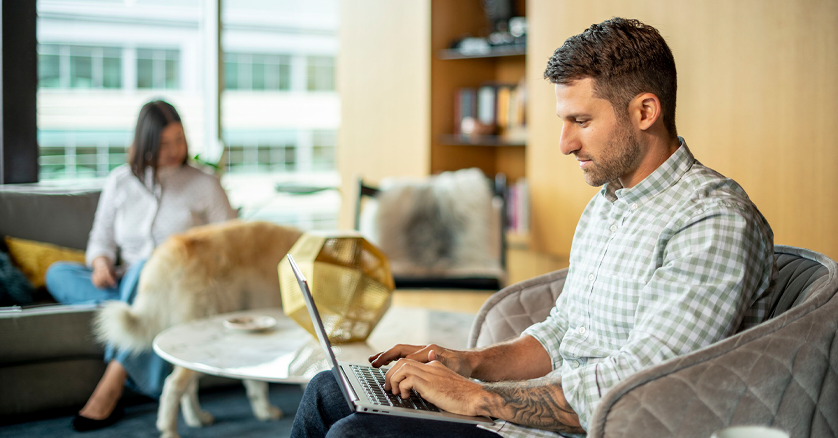 一名男子在家里用笔记本电脑工作，一名女子在背景中抚摸着一只狗